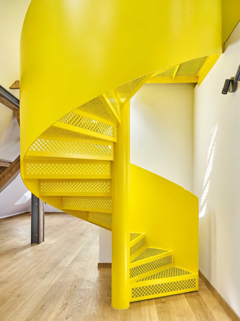 Eine knallig-gelbe Treppe aus Stahl mit kleinen, feinen Lasermotiven in den Tritt- und Setzstufen, die Transparenz und natürliche Lichteffekte schaffen, verbindet den Wohnraum in einem denkmalgeschützten Gebäude mit einer kleinen Galerie.